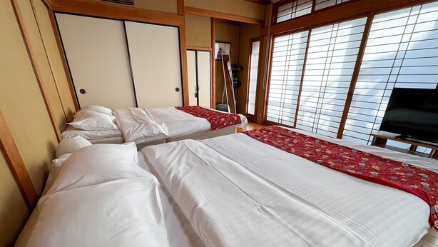 Habitación de hotel con dos camas, suelo de madera, televisión y ventanas amplias con persianas.