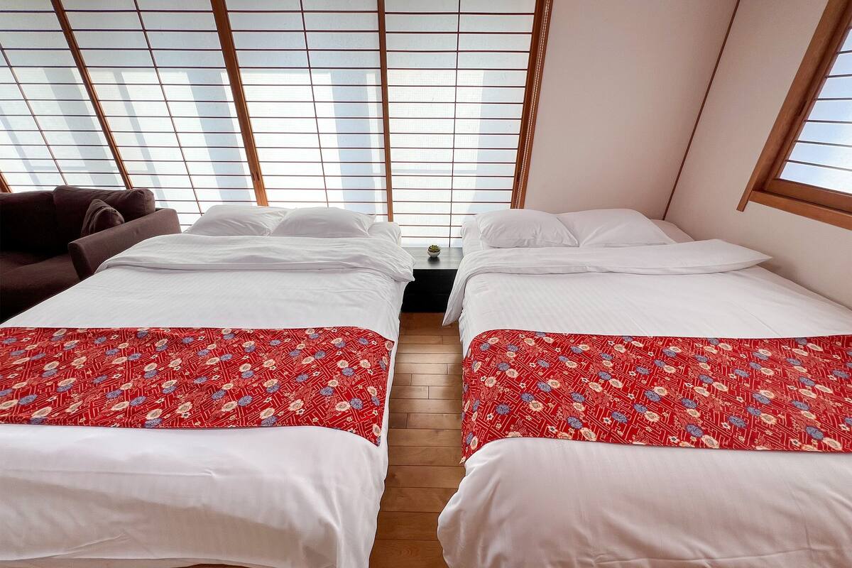 A room with two beds, each with a red patterned bedspread, white pillows, and a wooden floor.