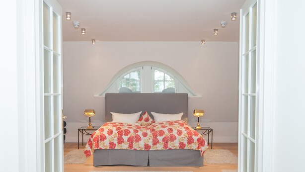A bedroom with a large bed, two bedside tables with lamps, a window with a curved top, and a white door. - Landhaus Töpferhof (Ratekau)