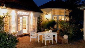 A well-lit outdoor patio with a table and chairs, surrounded by lush greenery and flowering plants. - Landhaus Töpferhof (Ratekau)