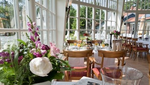 A sunlit dining area with large windows, wooden tables, and chairs. - Landhaus Töpferhof (Ratekau)