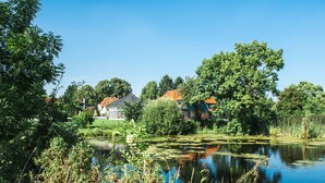 A serene lakeside setting with a house featuring a red roof and white walls, surrounded by lush greenery and trees. - Landhaus Töpferhof (Ratekau)