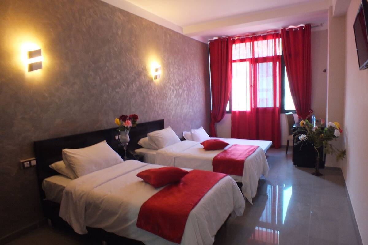 A hotel room with two beds, red and white bedding, a vase with flowers, and a red curtain.