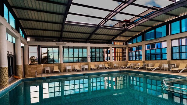 An indoor swimming pool with clear glass walls, lounge chairs, and a view of the outdoors. - Hotel Syphax (Bejaia)
