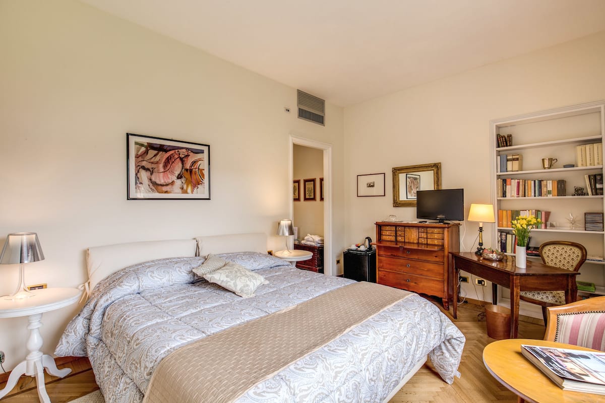 Ein Schlafzimmer mit einem großen Bett, Nachttischen, einem Schreibtisch mit Stuhl, einem Bücherregal und einem Fernseher.