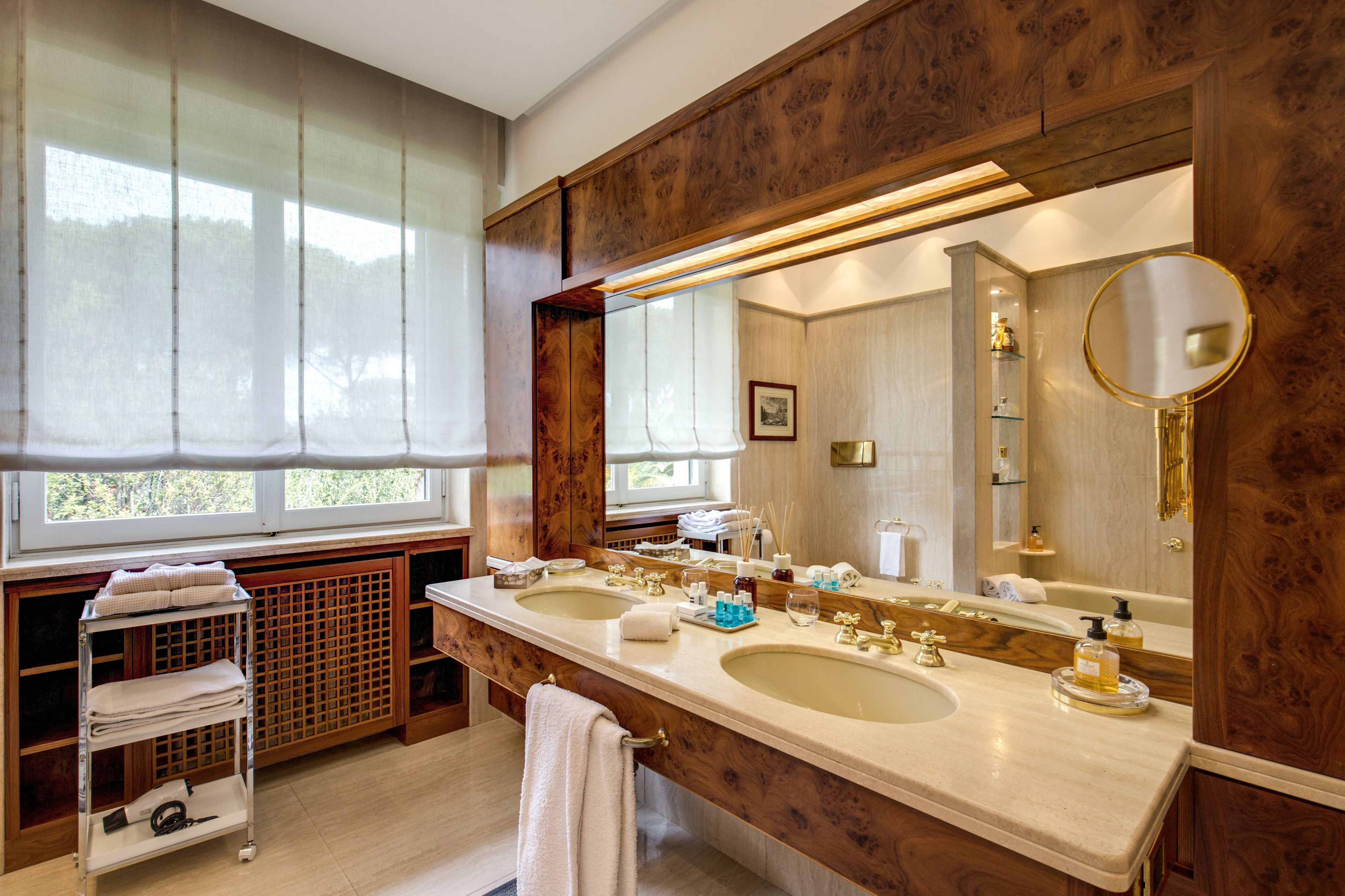 Un baño con dos lavamanos, un espejo grande, un perchero para toallas y una ventana con cortinas translúcidas.