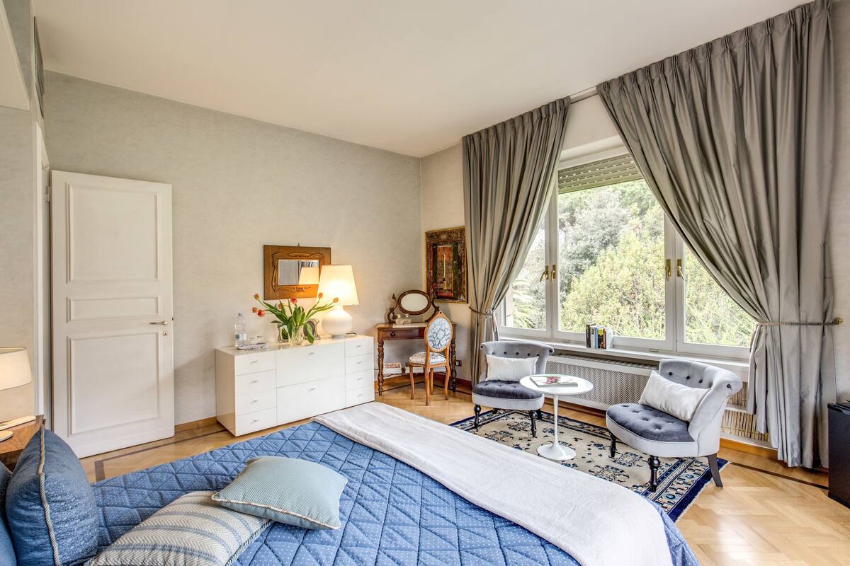Ein Schlafzimmer mit einem großen Bett, einem Schreibtisch, einem Stuhl, einem Heizkörper und einem Fenster mit Vorhängen.