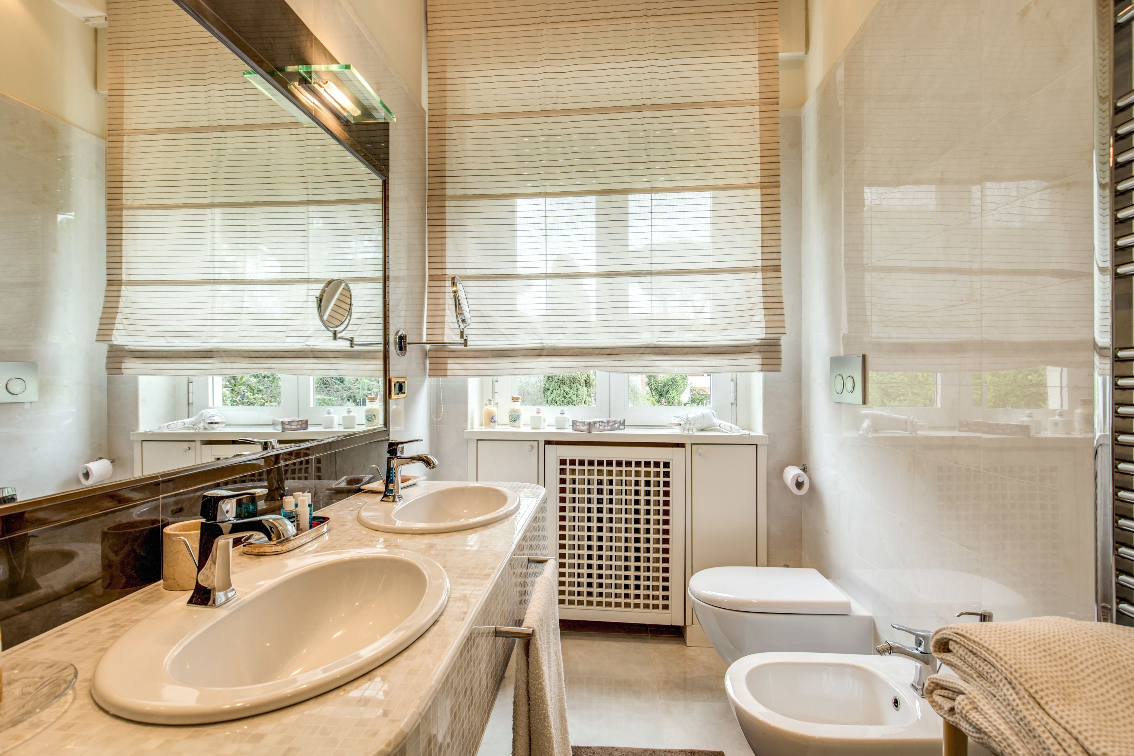 A modern bathroom with two sinks, a toilet, and a bidet. It features large windows with blinds, a mirror, and a towel rack.