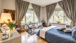 Un dormitorio con una cama grande, dos sillas, una mesita, una lámpara y una ventana con cortinas.