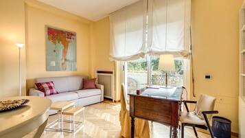 A living room with a sofa, a dining table, a chair, a lamp, and a window with curtains.