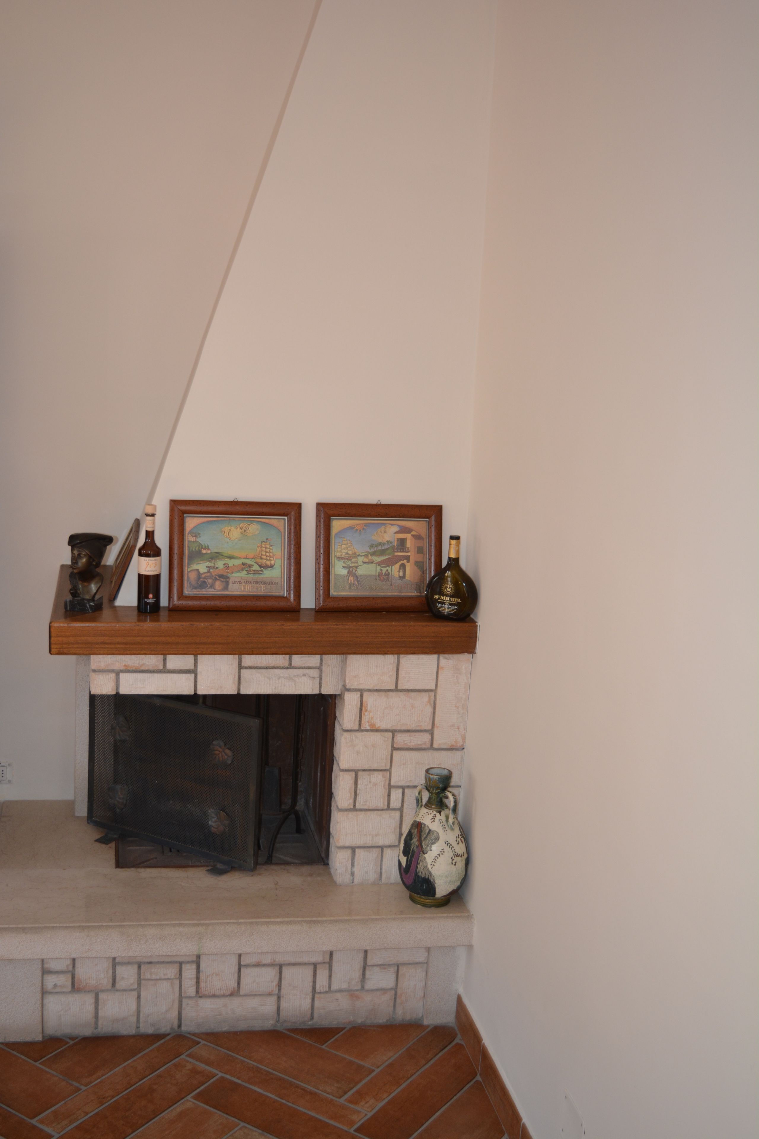 A fireplace with two framed artworks, a bottle, and a vase on the mantel.