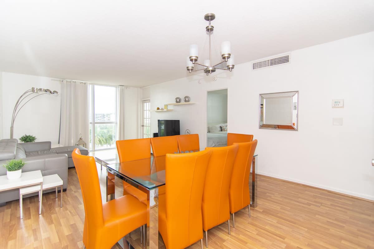 A modern living room with a dining area, featuring a sofa, orange chairs, a glass table, and a chandelier.