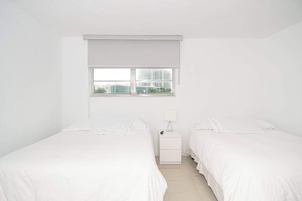 A minimalist bedroom with two beds, a nightstand, and a window with a view of buildings.