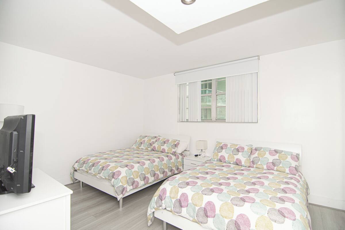 A bedroom with two beds, a TV, and a window with blinds.