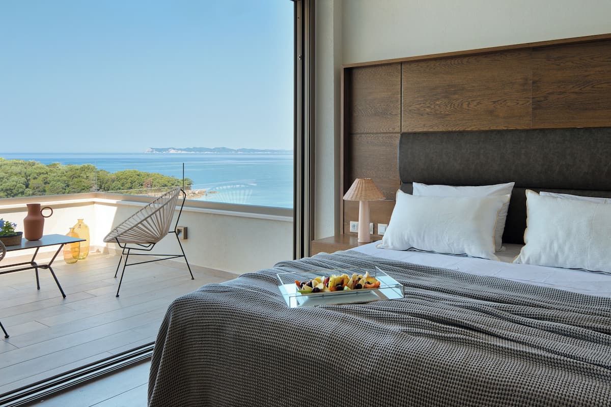 Habitación de hotel con una cama grande, un balcón y vistas al mar, además de una mesa con una bandeja de frutas.