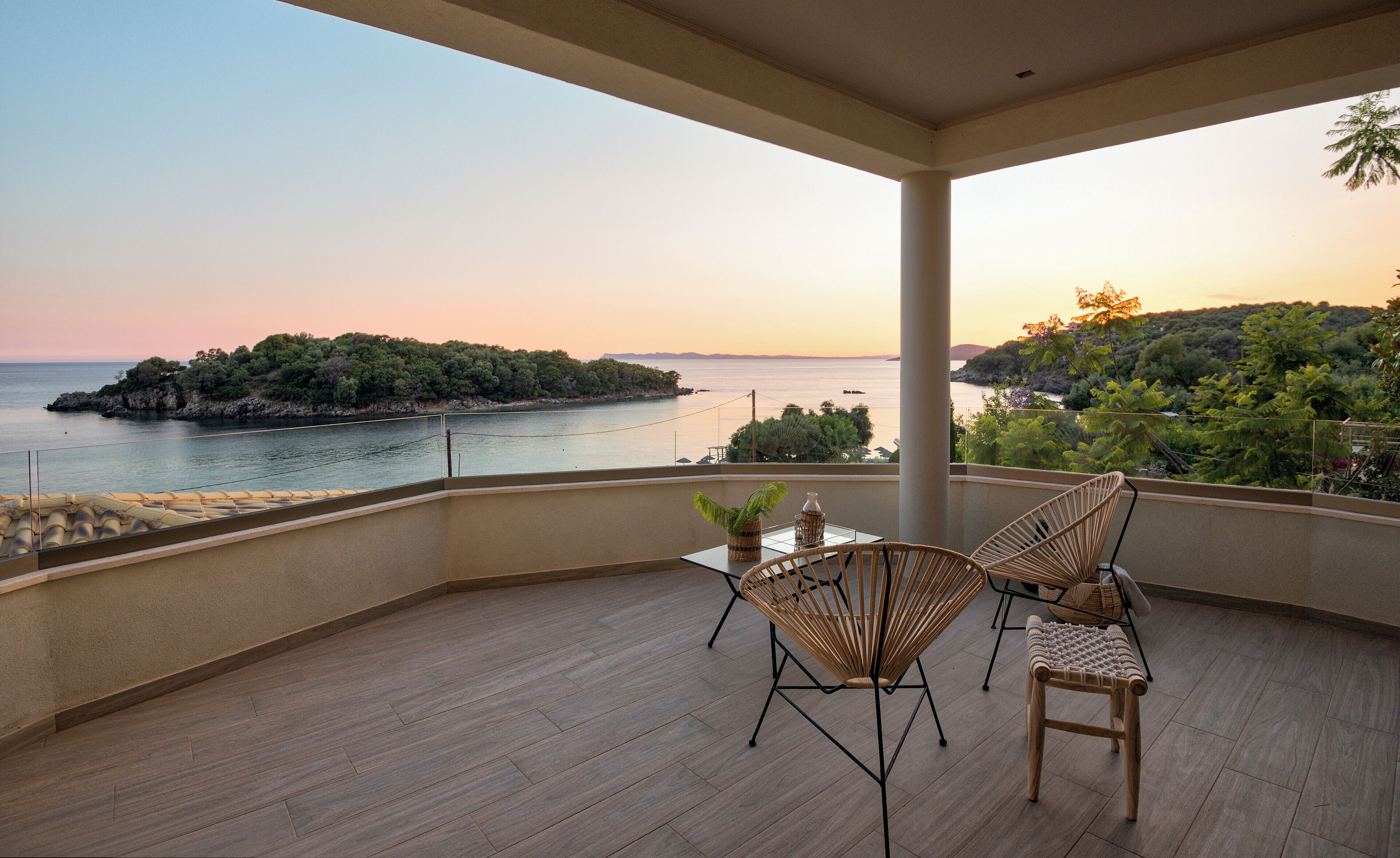 A balcony with two wicker chairs, a small table, and a view of the sea and a distant island at sunset.