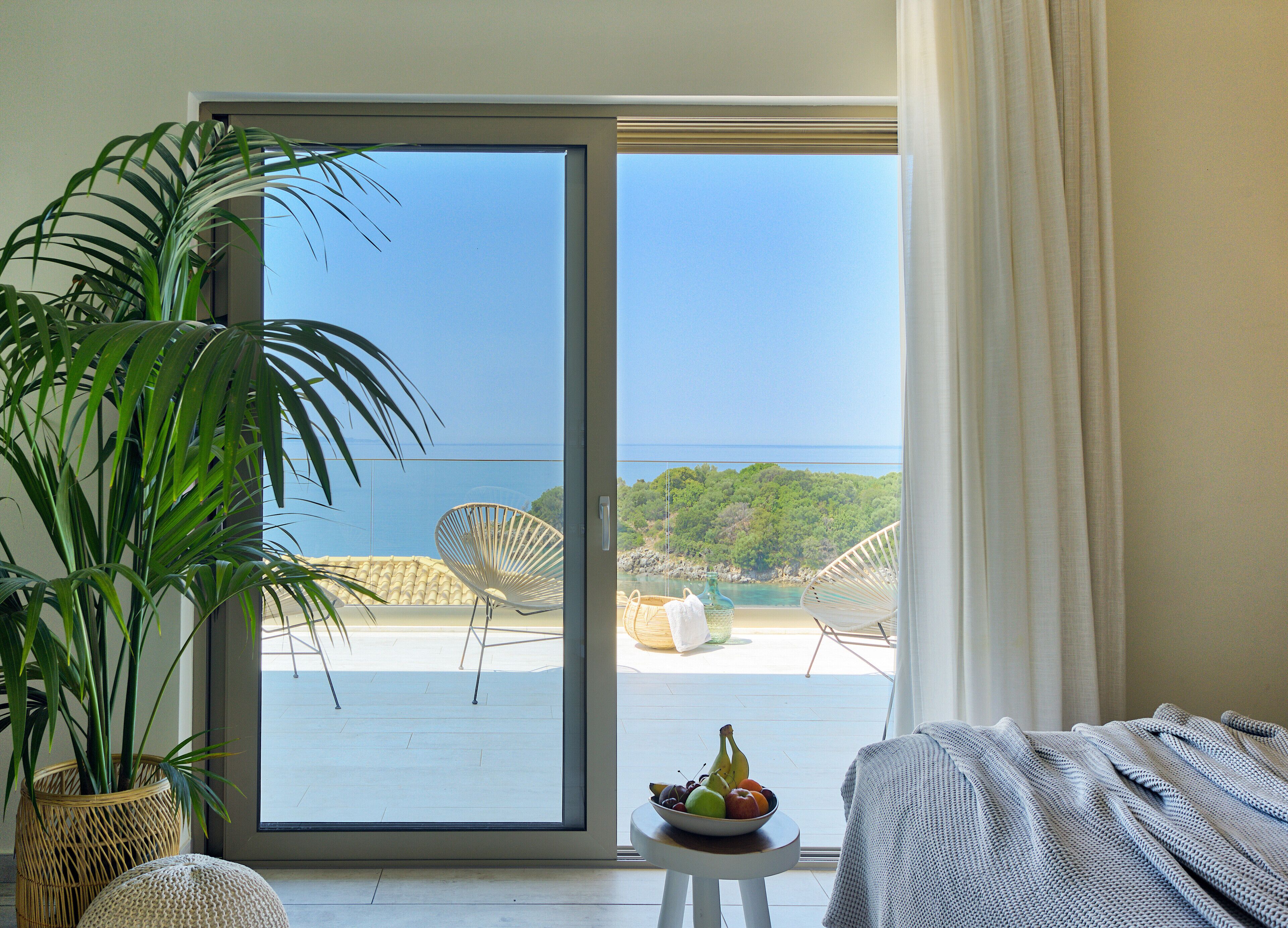Une pièce dotée d’une grande porte-fenêtre coulissante donnant sur un balcon avec vue sur la mer et la végétation. On y trouve une chaise en osier et une petite table sur laquelle est posé un saladier rempli de fruits.