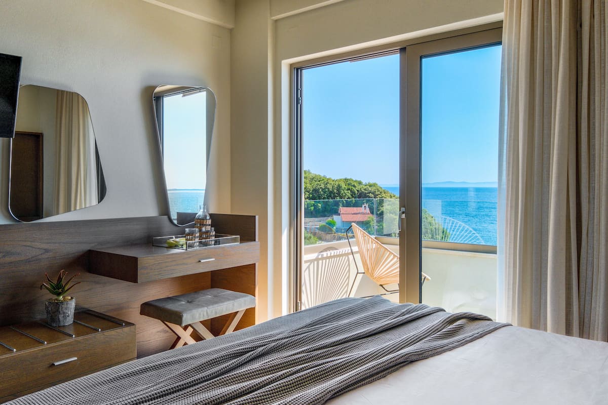 Un dormitorio con una cama grande, una mesita de noche, un espejo y vistas al mar a través de un ventanal corredero.