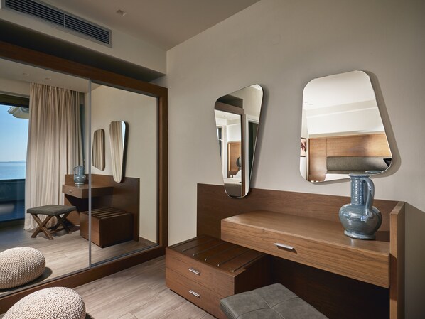 A modern hotel room with a wooden vanity, a large mirror, a bedside table with a lamp, a small stool, and a view of the sea through a large window.