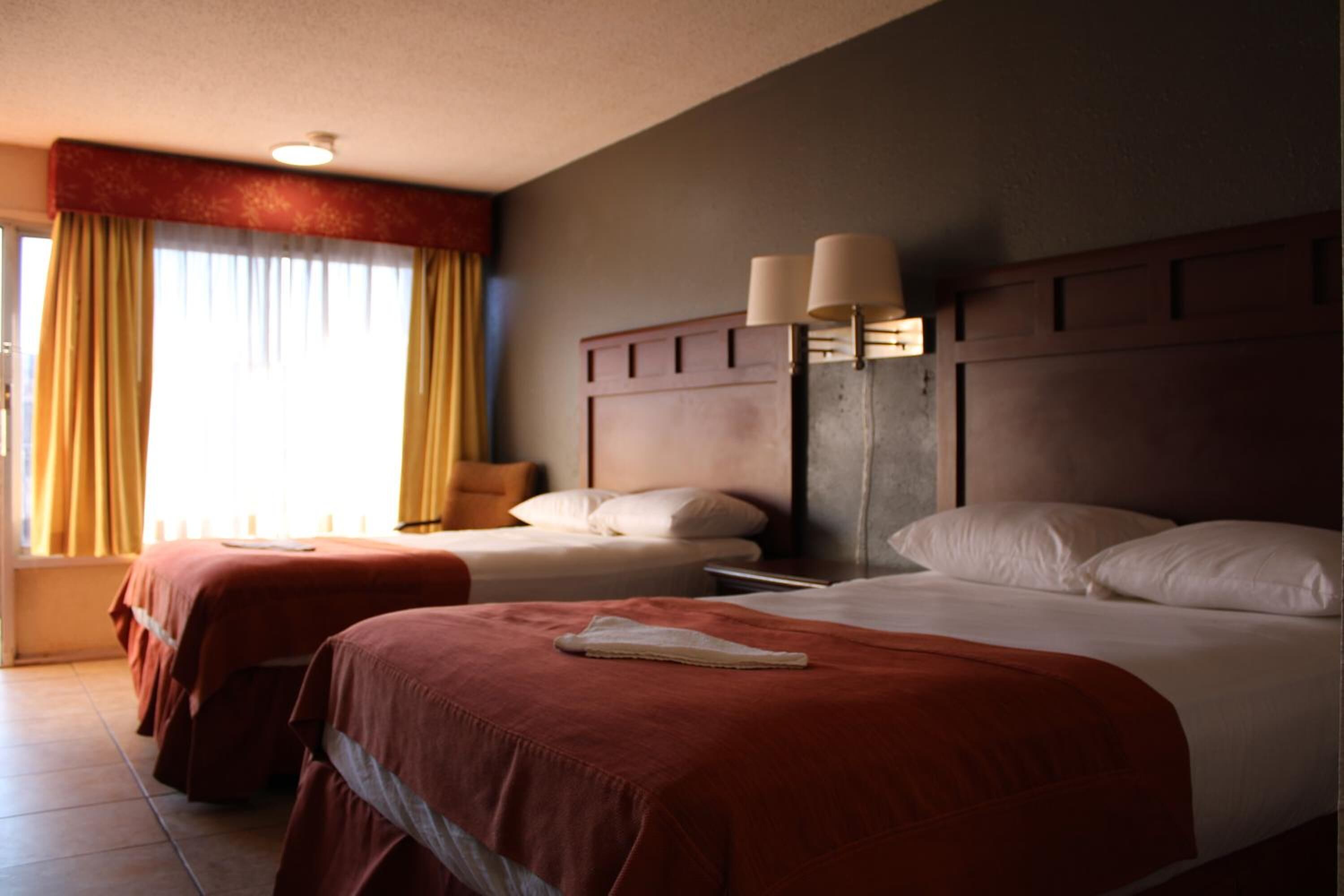 A hotel room with two beds, a nightstand, a lamp, and a window with curtains.