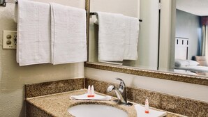 A bathroom with a granite countertop sink, two toothbrushes, and a mirror reflecting a bed and a nightstand.