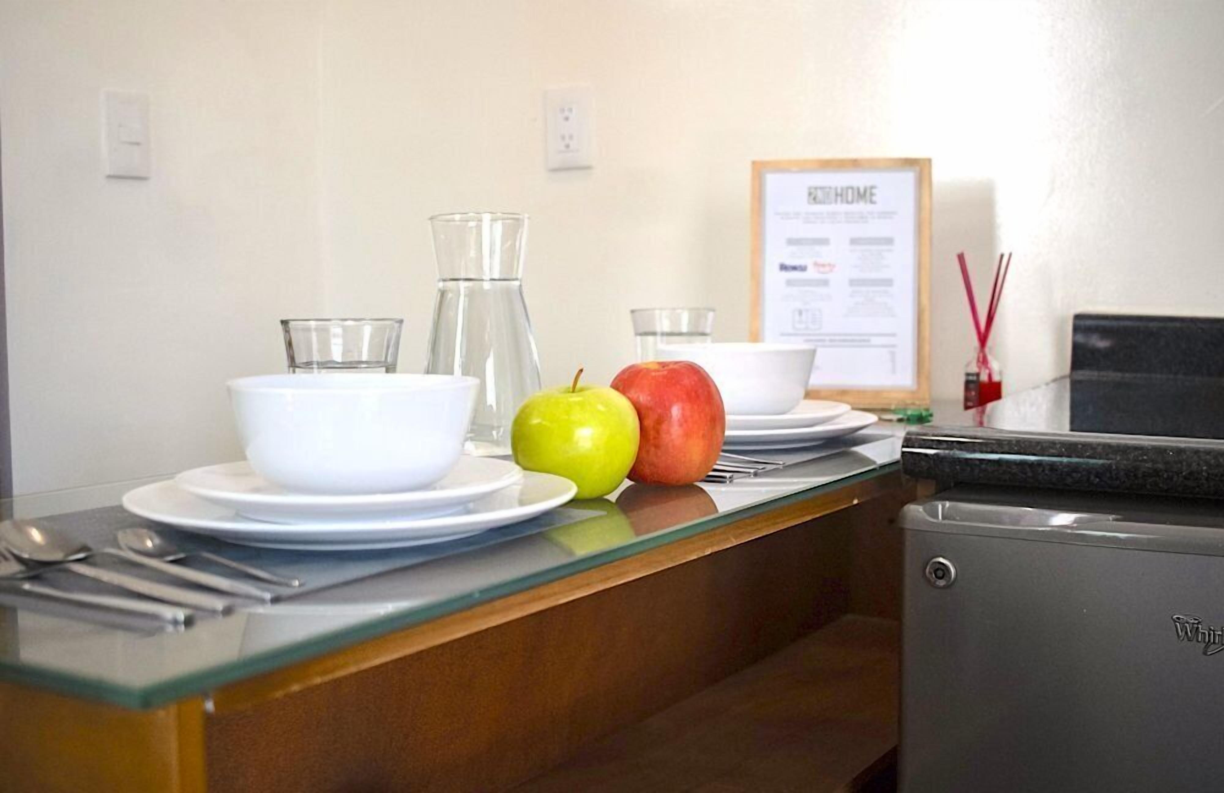 Una encimera de cocina con un refrigerador, una jarra de vidrio, un tazón, un plato, una cuchara, un tenedor y dos manzanas.