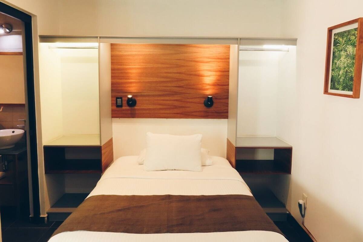 Una habitación de hotel con cama, mesitas de noche, un espejo y una lámpara fijada en la pared.