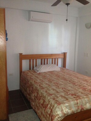 A bedroom with a wooden bed frame, a floral bedspread, a white pillow, a ceiling fan, and an air conditioning unit.