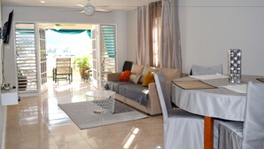 A modern living room with a sofa, dining table, and chairs. There is a TV mounted on the wall, a ceiling fan, and a view of the outdoors through a door. - Bay Pointe Yacht Club 2BD Apartment OLR (Montego Bay)