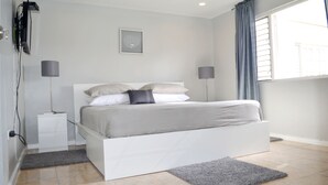 A modern bedroom with a white bed, grey bedding, a small bedside table, a floor lamp, and a window with curtains. - Bay Pointe Yacht Club 2BD Apartment OLR (Montego Bay)