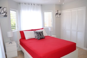 A bedroom with a red bedspread, white pillows, a bedside table with a lamp, and a window with blinds.