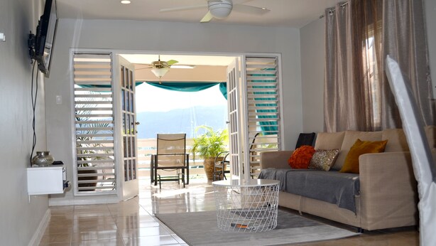 A living room with a sofa, a coffee table, a TV mounted on the wall, and a view of the outdoors through an open door. - Bay Pointe Yacht Club 2BD Apartment OLR (Montego Bay)
