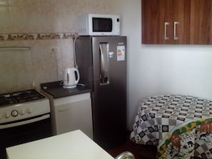 Cocina compacta con estufa, microondas, hervidor, refrigerador y una mesa con mantel estampado.