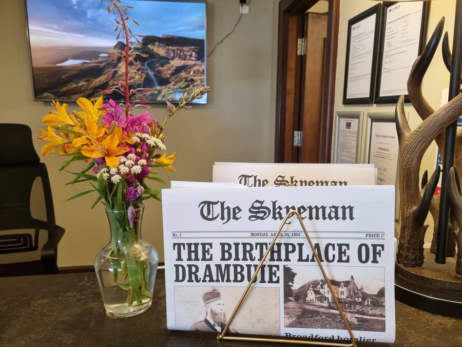A newspaper titled "The Skewman" with a headline about Drambuie, a glass vase with flowers, and a framed picture of a landscape.