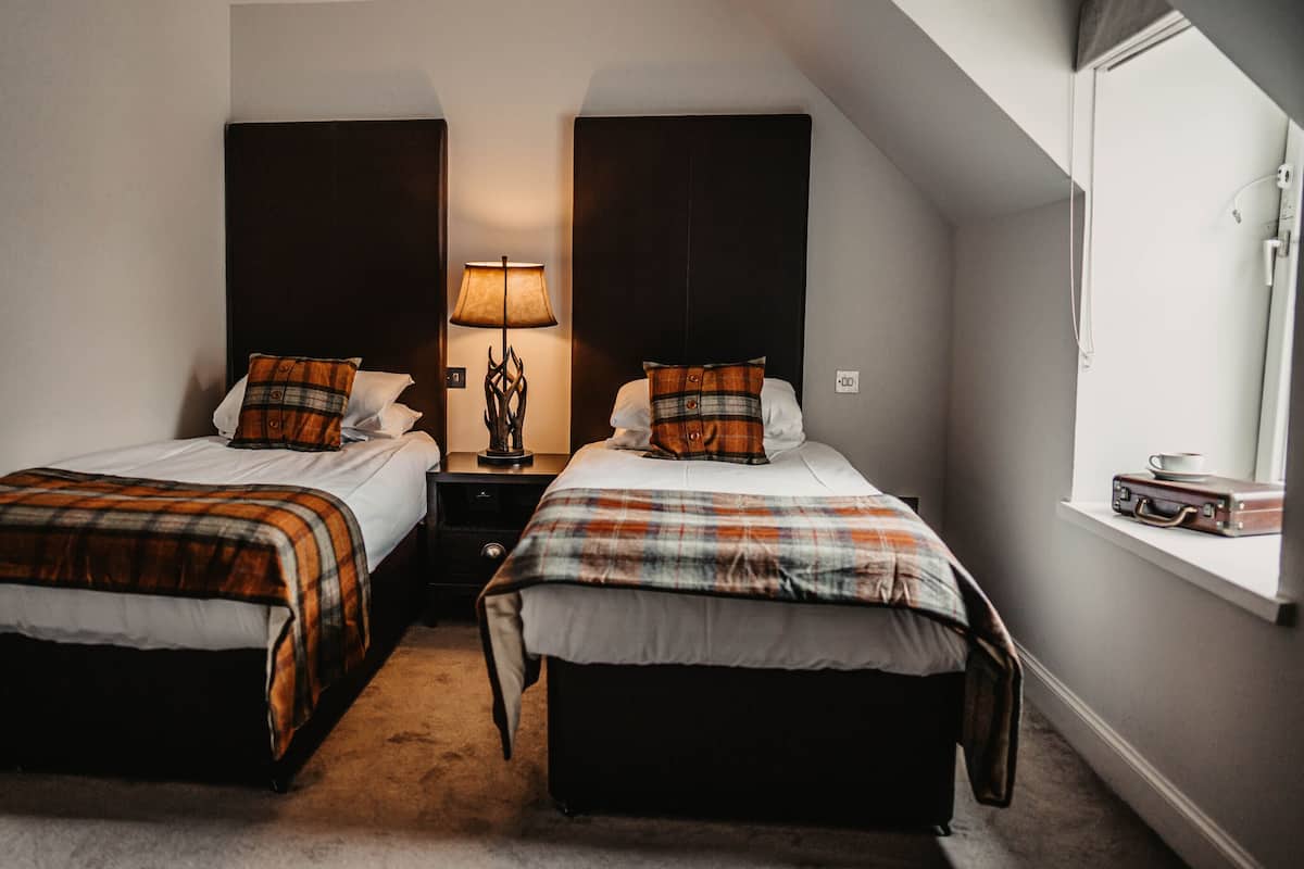 A room with two beds, a nightstand, a lamp, and a suitcase on a windowsill.