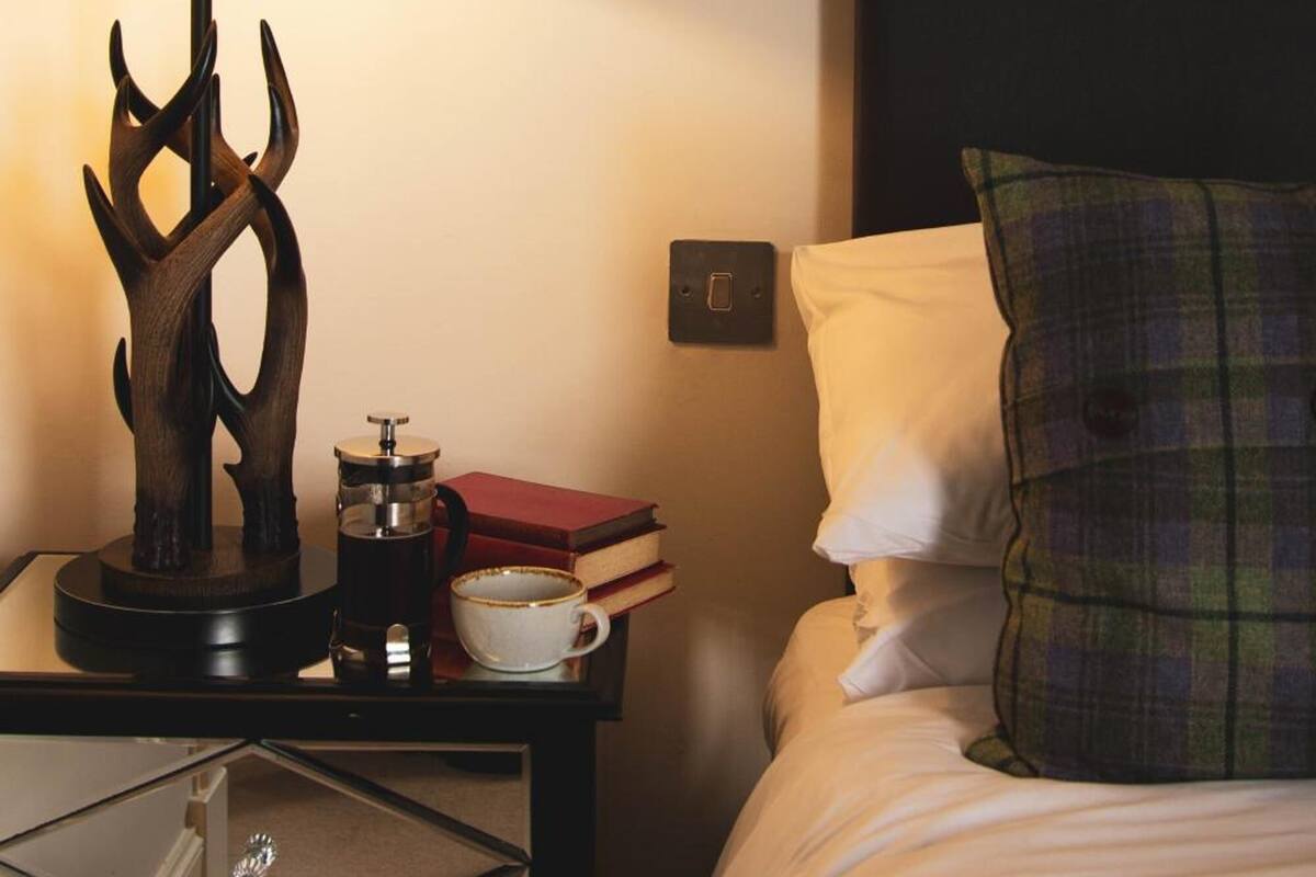 A neatly made bed with a plaid pillow, a bedside table with a lamp, a coffee maker, and a book.