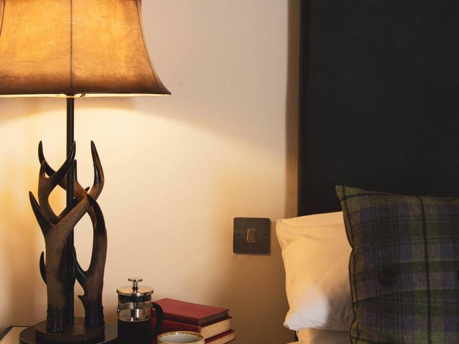 A neatly made bed with a plaid pillow, a bedside table with a lamp, a coffee maker, and a book.