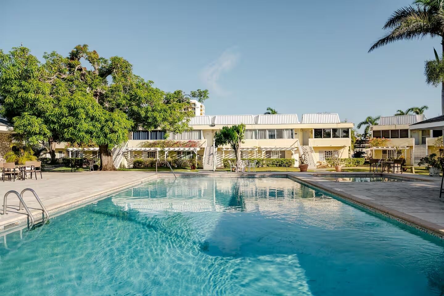 Una piscina con superficie de agua azul cristalina, rodeada por un patio con mobiliario exterior y un edificio de fachada blanca al fondo.