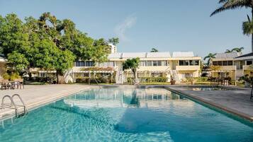 Una piscina con superficie de agua azul cristalina, rodeada por un patio con mobiliario exterior y un edificio de fachada blanca al fondo.