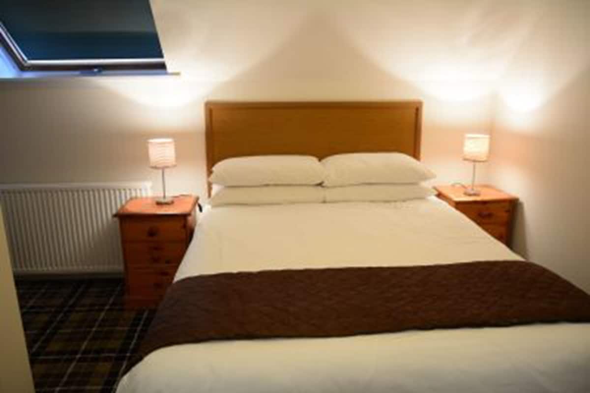 Ein ordentlich bezogenes Bett mit Holz-Kopfteil in einem Hotelzimmer. Zwei Nachttische mit Lampen sind vorhanden, und an der Wand ist ein Heizkörper zu sehen.