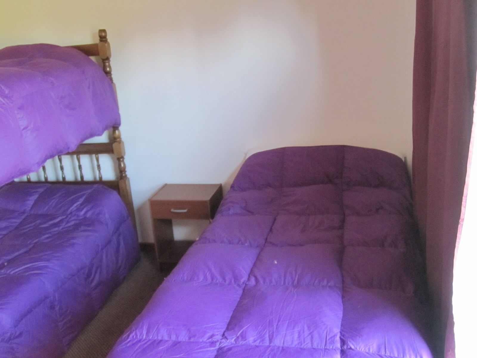 A room with two single beds, a wooden headboard, a small wooden nightstand, and a purple blanket.