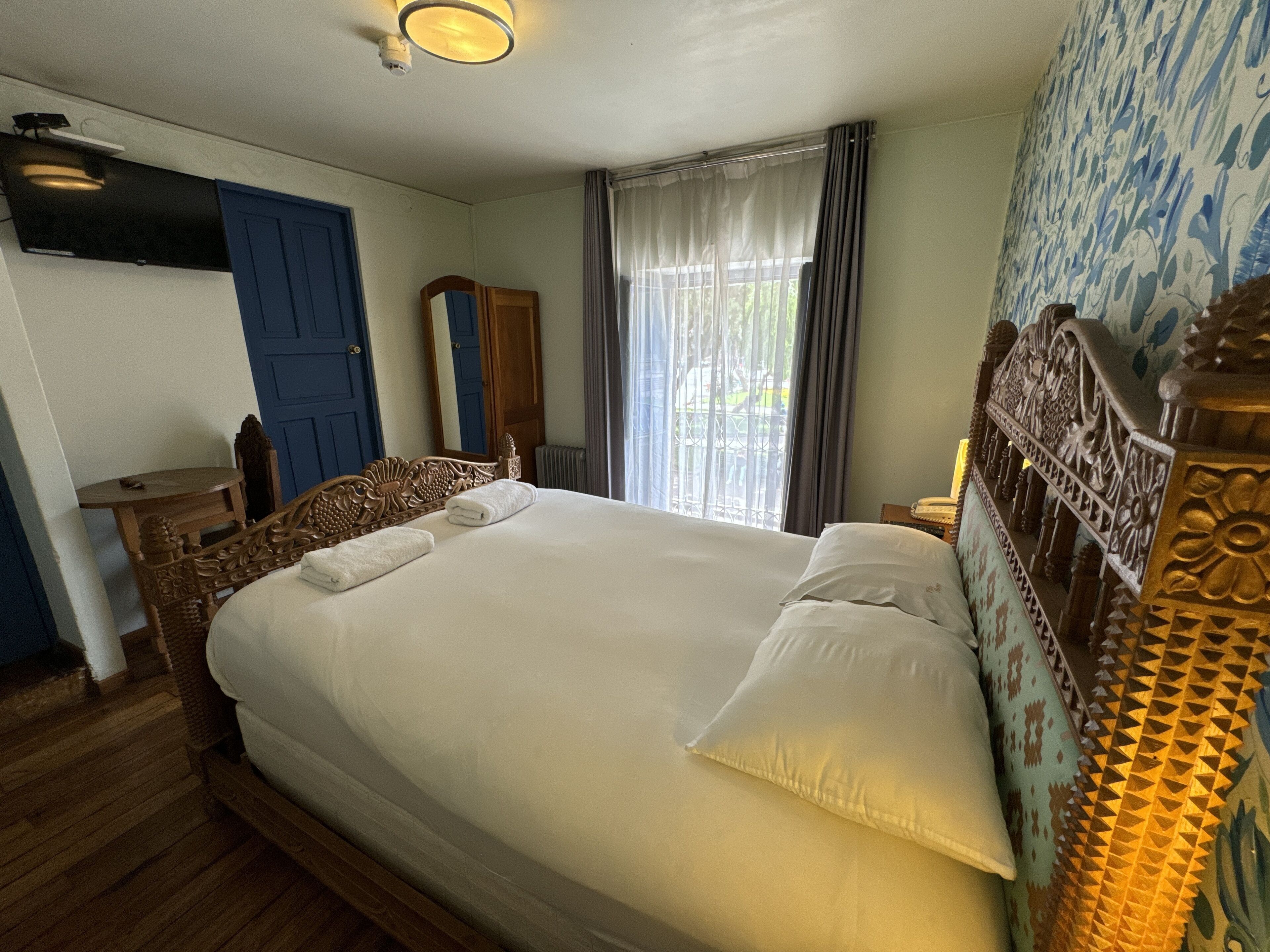 A hotel room with a large bed, a wooden headboard, a small table with a chair, a mirror, and a window with curtains.