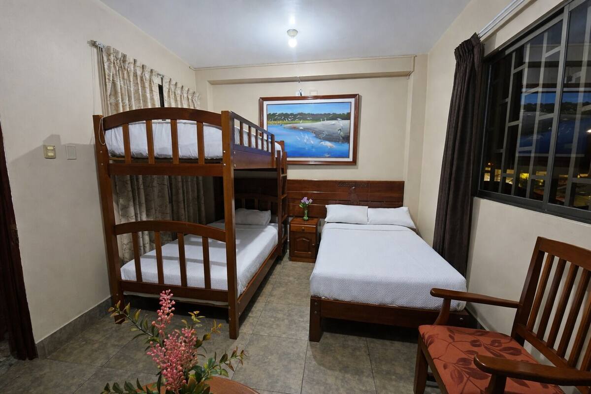 Una habitación con literas, una cama individual, una mesita de noche con un florero, una silla y una ventana con cortinas.