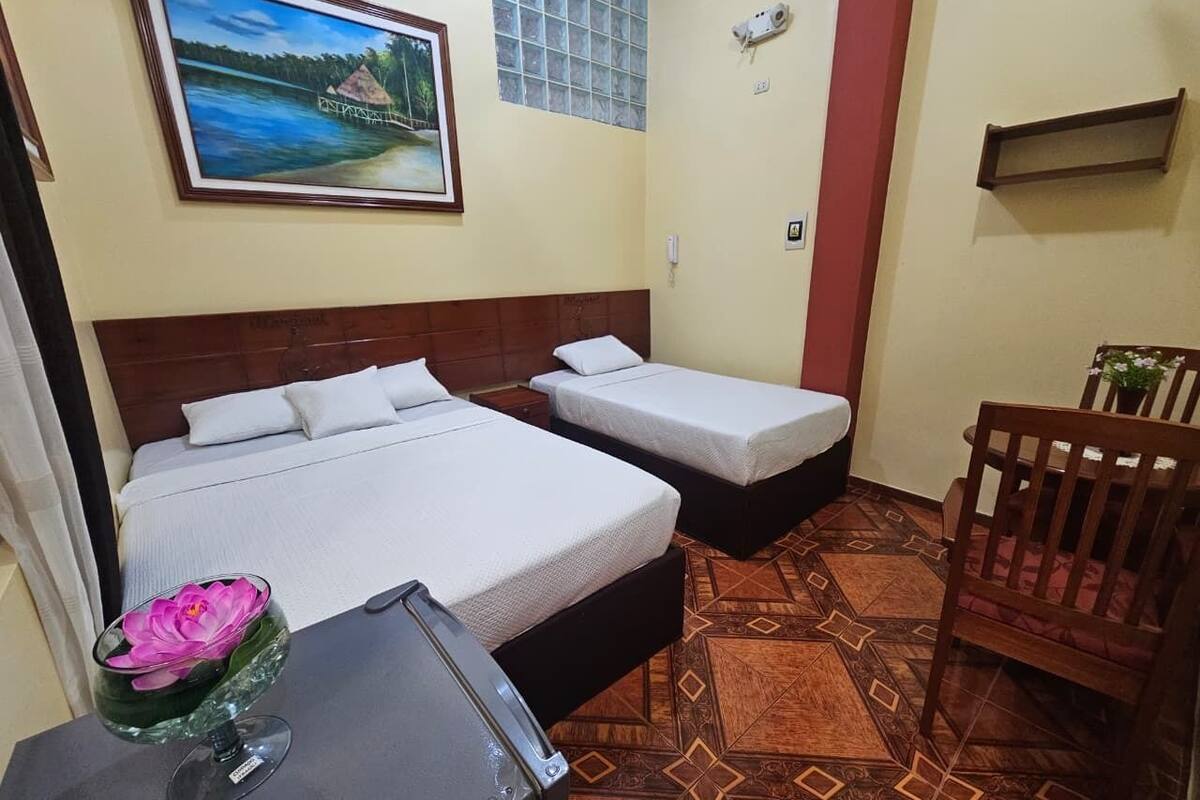 Una habitación con dos camas, una mesita con una flor, una silla, un cuadro en la pared y un suelo estampado.