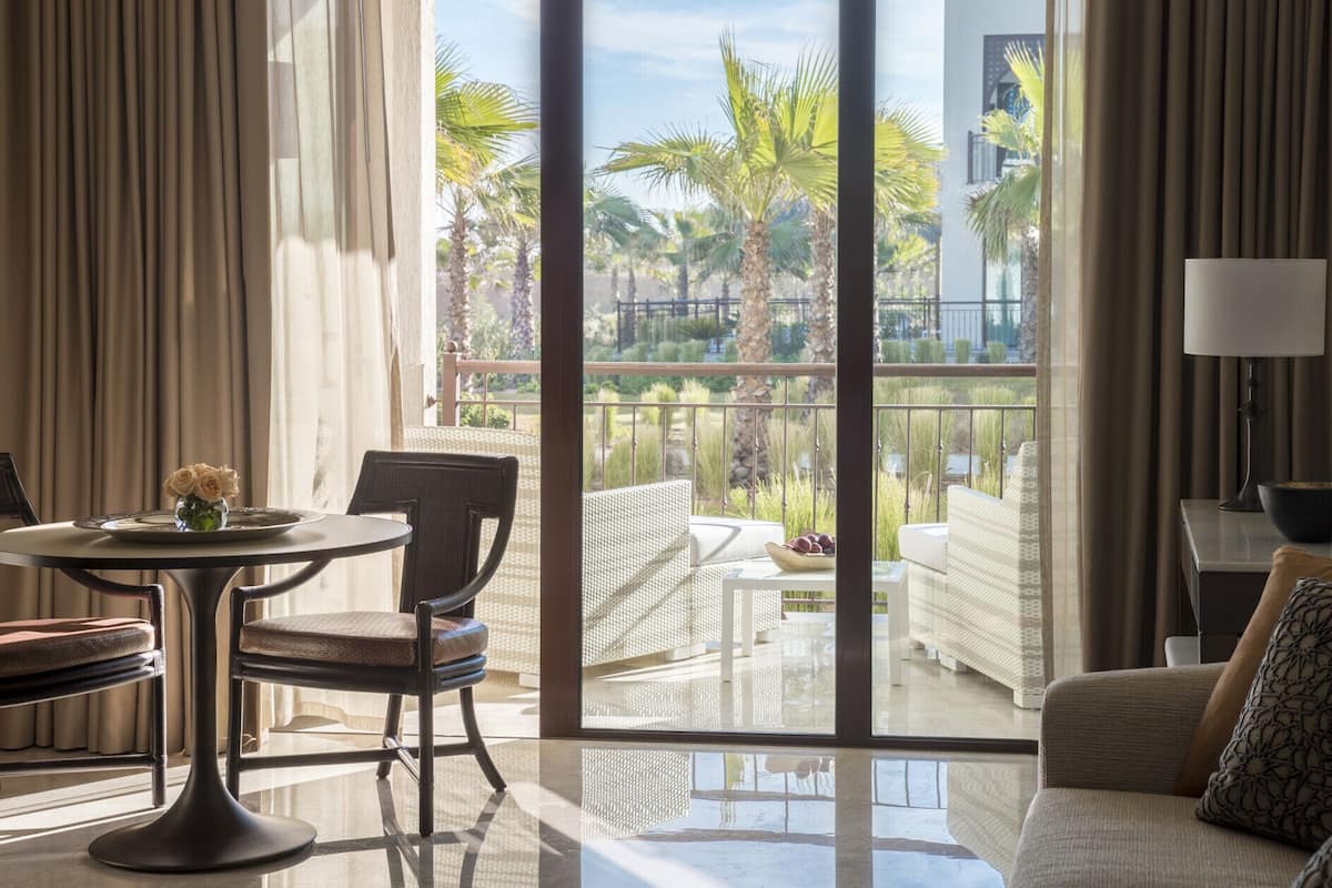 Quarto espaçoso com uma grande porta de vidro deslizante que dá para uma varanda com vista para palmeiras e uma piscina. Há uma mesa redonda com cadeiras e um sofá.