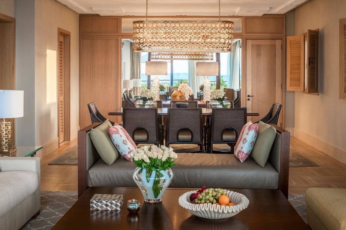 Uma sala de jantar moderna com um candelabro, um sofá com almofadas, uma mesa com uma cesta de frutas e vista para o exterior.