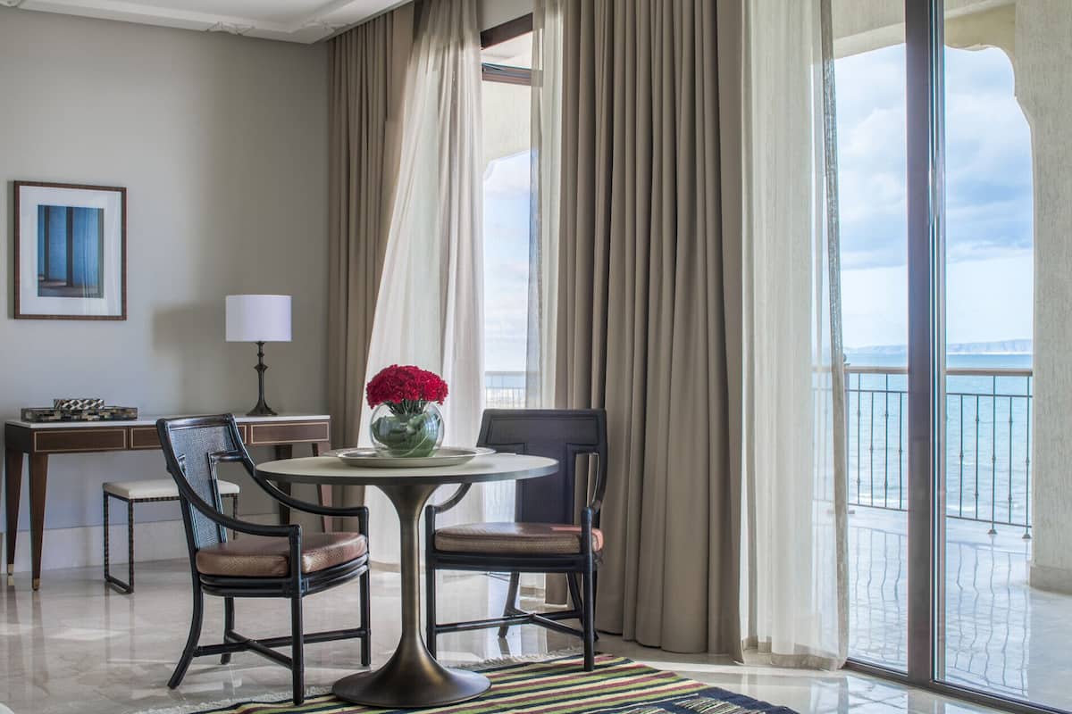 Quarto de hotel com mesa redonda, duas cadeiras, um vaso com flores vermelhas, abajur, quadro pendurado na parede e vista para o mar através de grandes janelas.