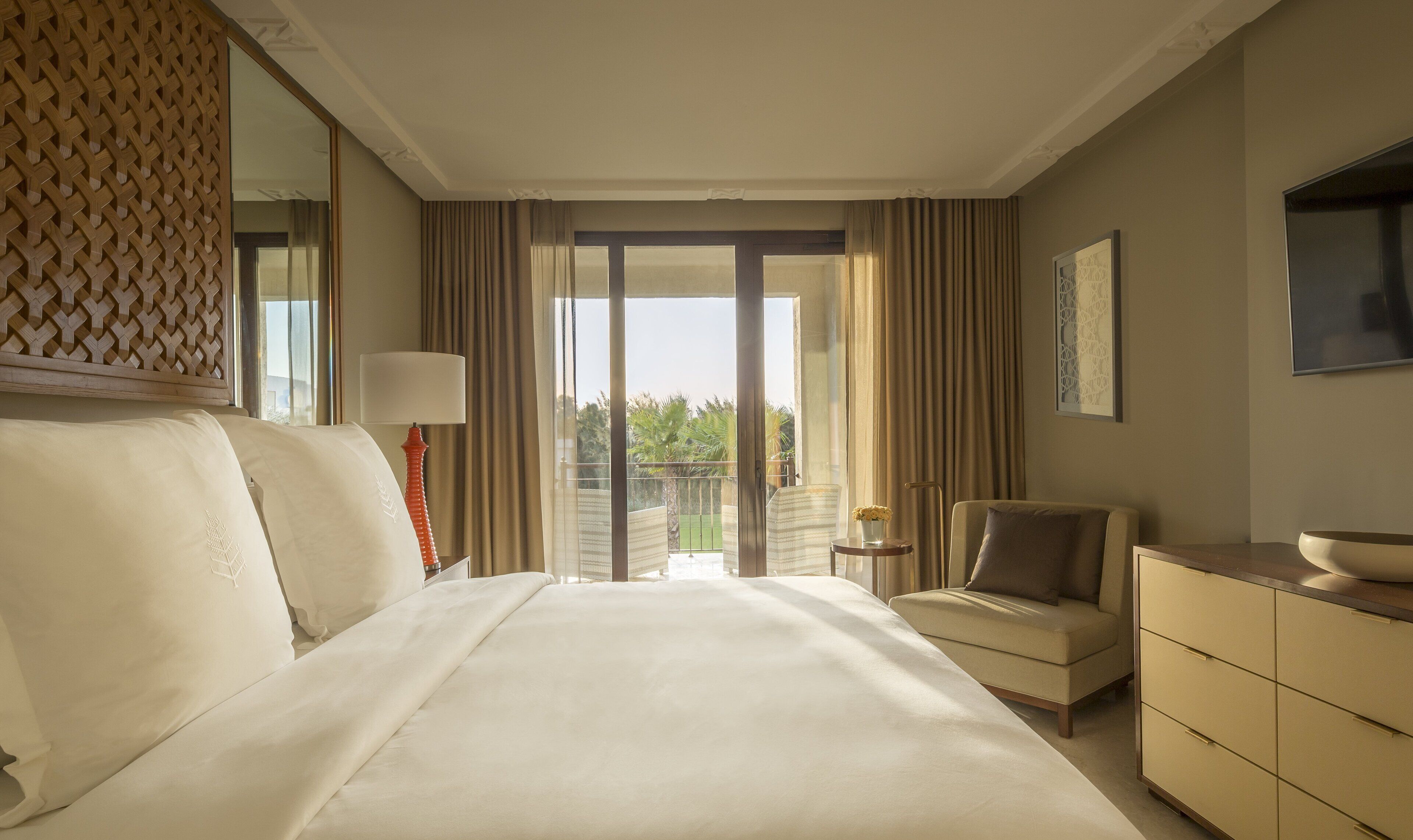 Habitación de hotel con una cama grande, un escritorio, una silla, un televisor y un balcón con cortinas.