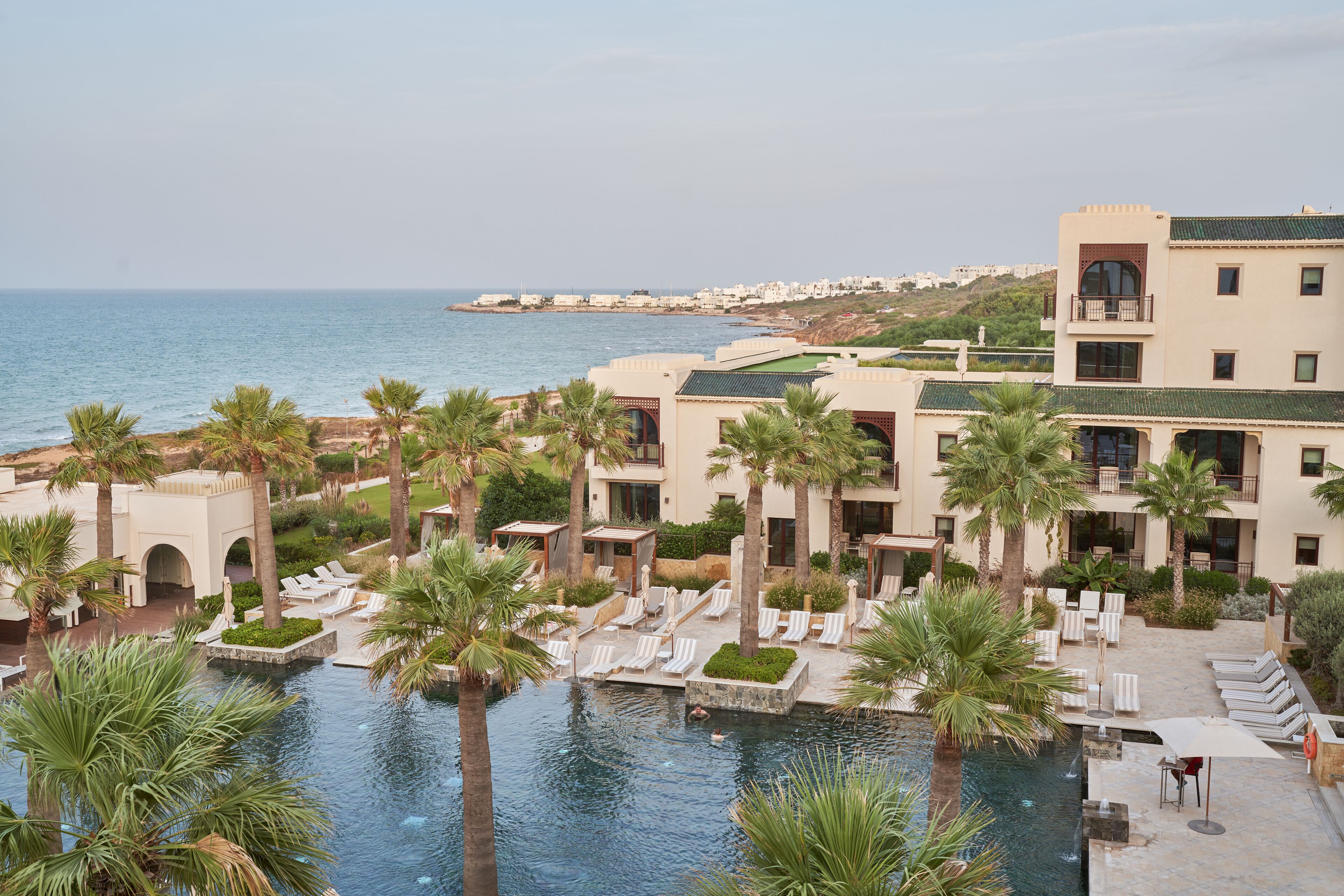 Un resort con piscina rodeada de palmeras y sillas de descanso, con vista al mar.