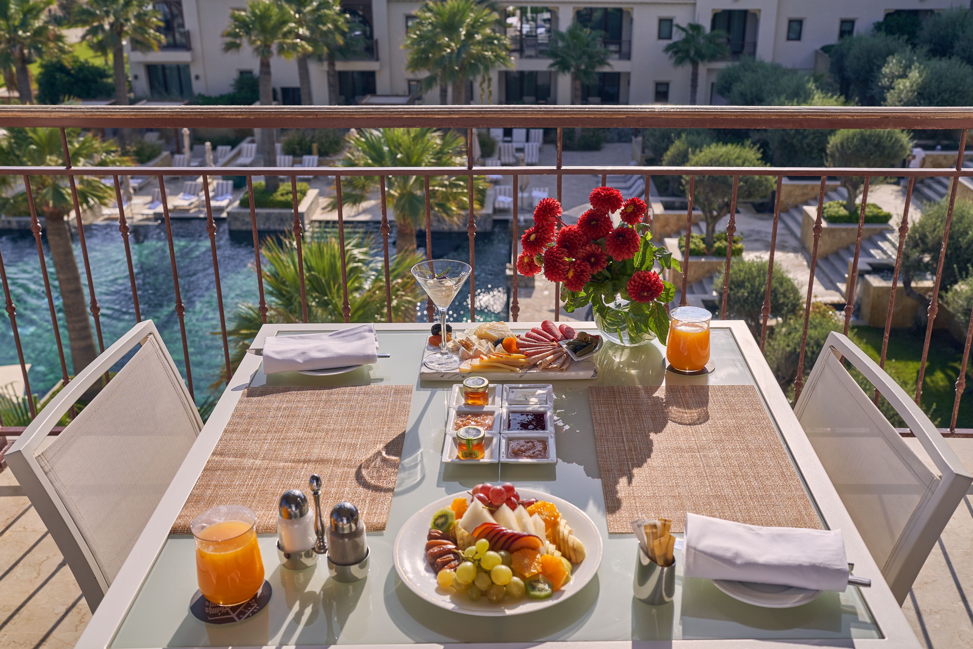 Una mesa de comedor con una bandeja de frutas, una copa de martini y un florero con flores rojas en un balcón con vista a una piscina y palmeras.
